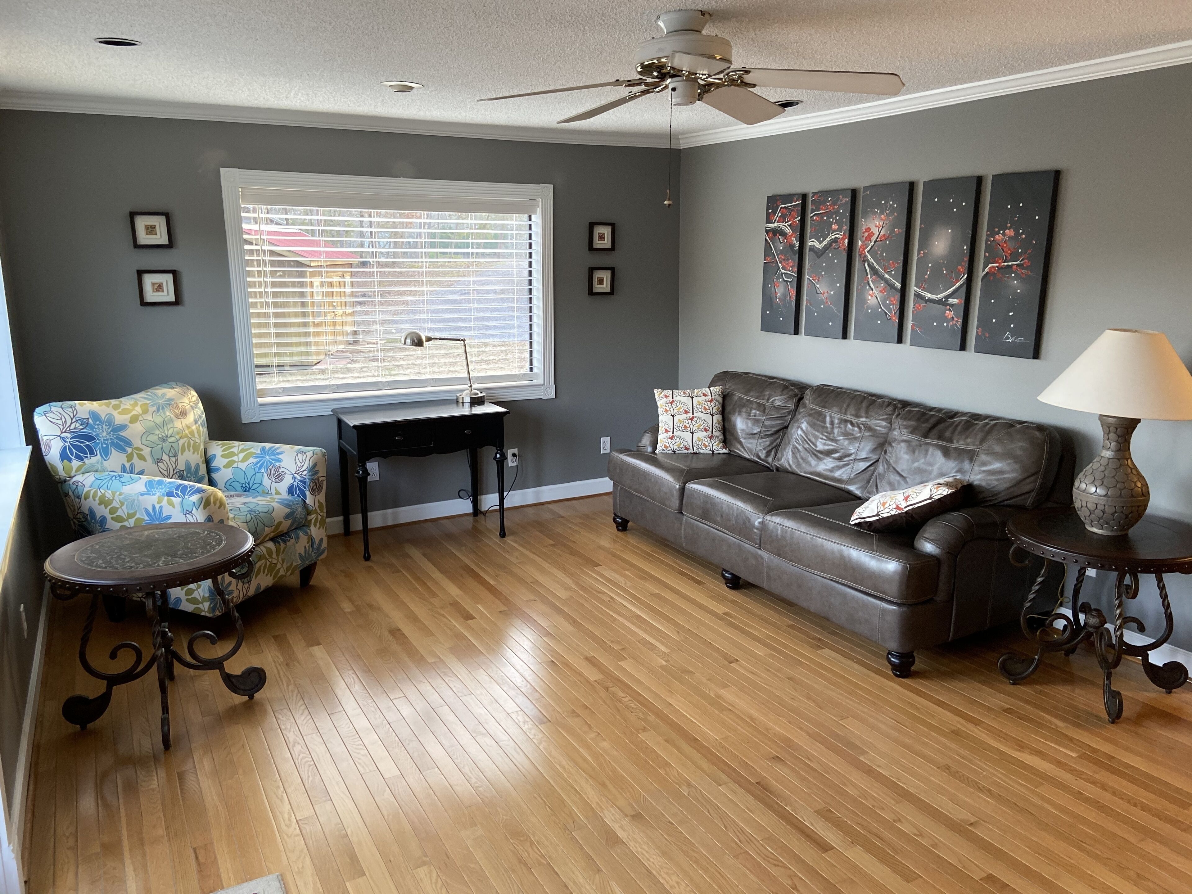 Un salon comprenant un canapé en cuir, un fauteuil à motifs floraux, une table basse en bois, une table d’appoint avec une lampe et un ventilateur de plafond.