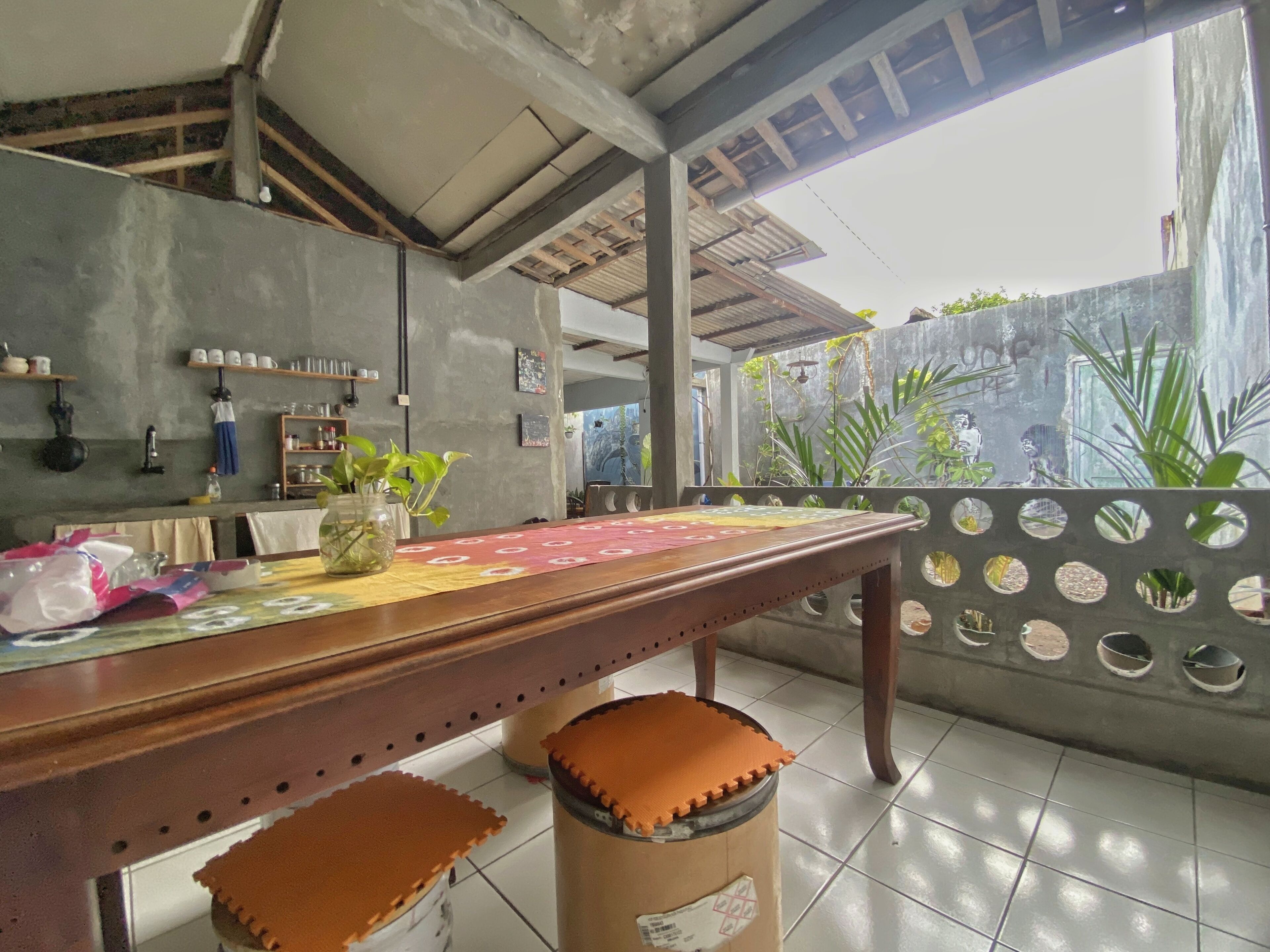 A kitchen area with a wooden table, stools, and a tiled floor. There are shelves with kitchen items and a window with plants outside.