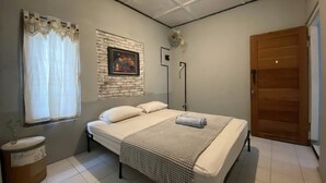 A bedroom with a bed, a window with curtains, a wall-mounted picture, a bedside table, and a door. - Reene'o Guesthouse (Yogyakarta)