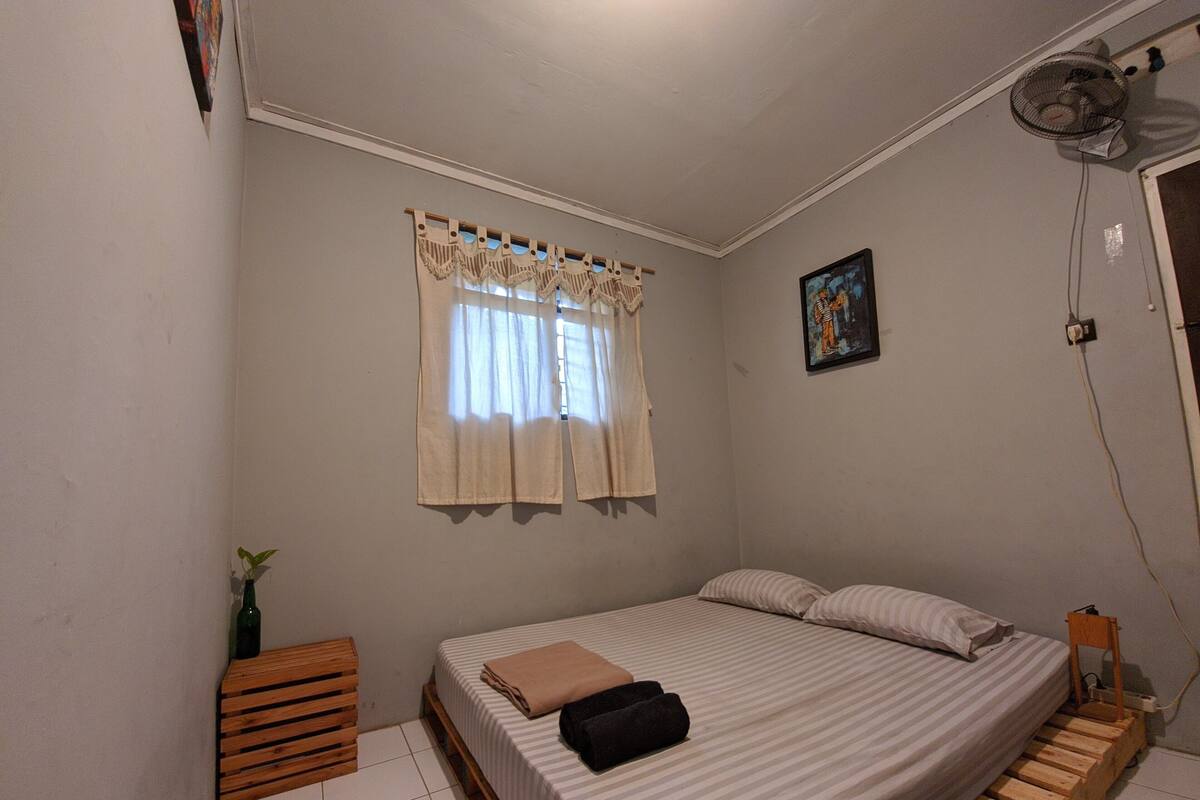 A bedroom with a bed, a window with curtains, a small bedside table, and a wall-mounted fan.