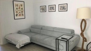 Ein modernes Wohnzimmer mit einem grauen Sofa, einem Glastisch und einer Lampe. An den Wänden befinden sich botanische Drucke in Bilderrahmen.