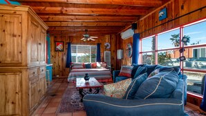A cozy cabin interior with wooden walls, a large window, a ceiling fan, and a bed with patterned bedding. - Chucks cabin Joshua Tree region, your desert Oasis (TWENTYNIN PLAMs)