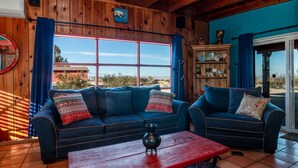 A cozy living room with two blue sofas, a wooden coffee table, and a large window with blue curtains. - Chucks cabin Joshua Tree region, your desert Oasis (TWENTYNIN PLAMs)