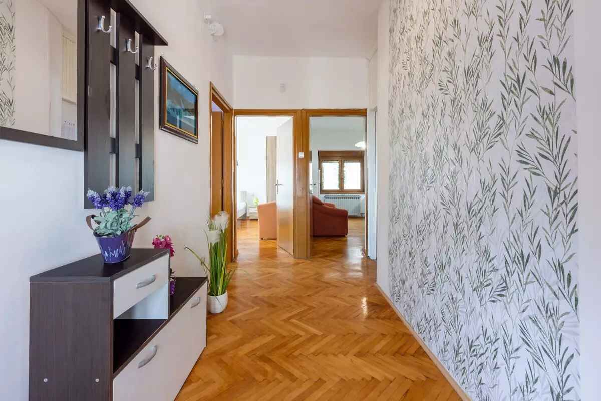 Un pasillo con piso de madera, un mueble de madera oscura con cajones, un espejo y una pared con estampado de hojas.