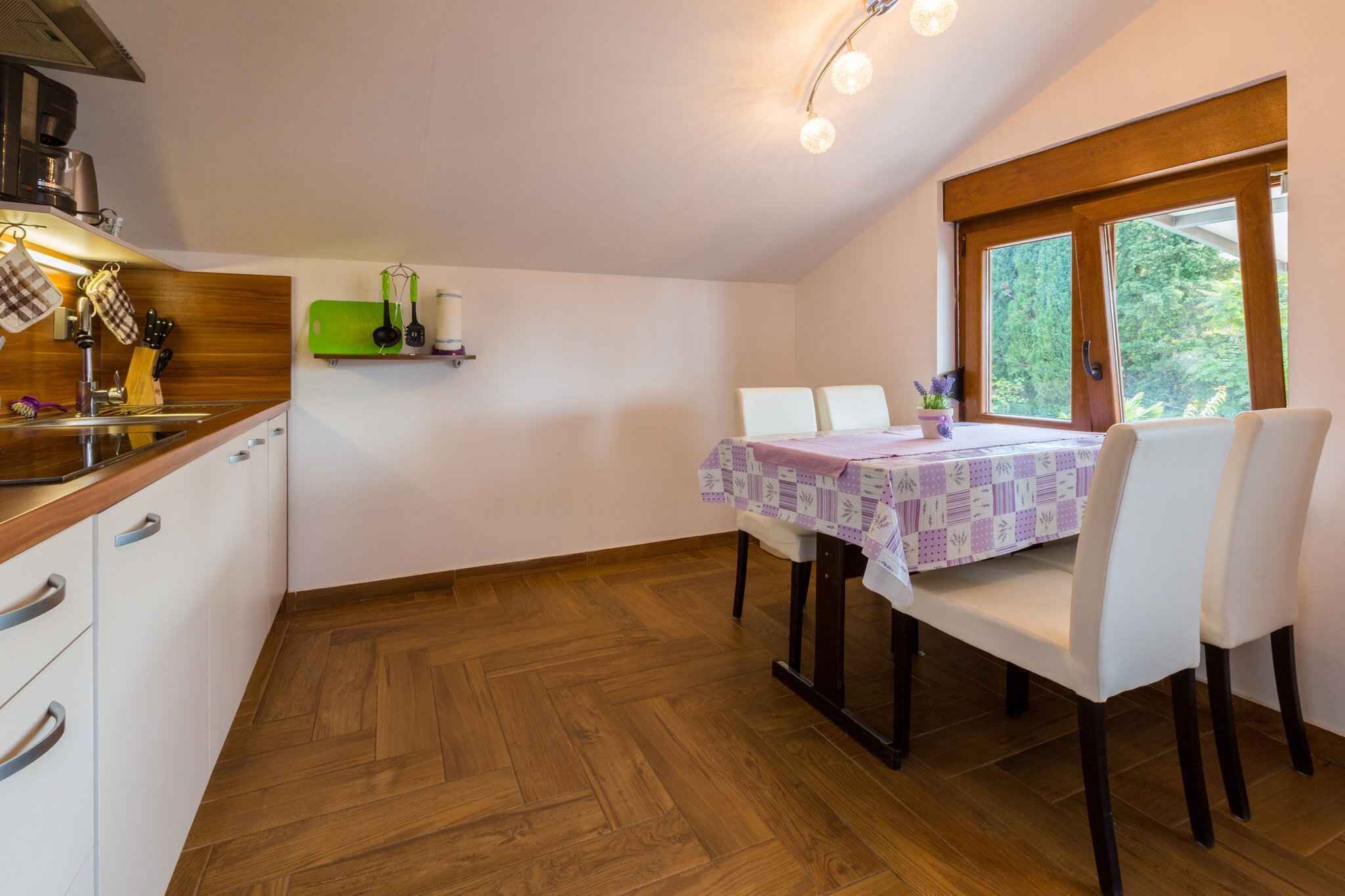 Eine kompakte Küche mit Holzparkett, einem Esstisch mit weißen Stühlen und einem Fenster mit Blick auf Grünflächen.