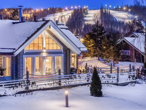 Un albergue cubierto de nieve con un amplio ventanal, una terraza de madera y una pista de esquí al fondo.