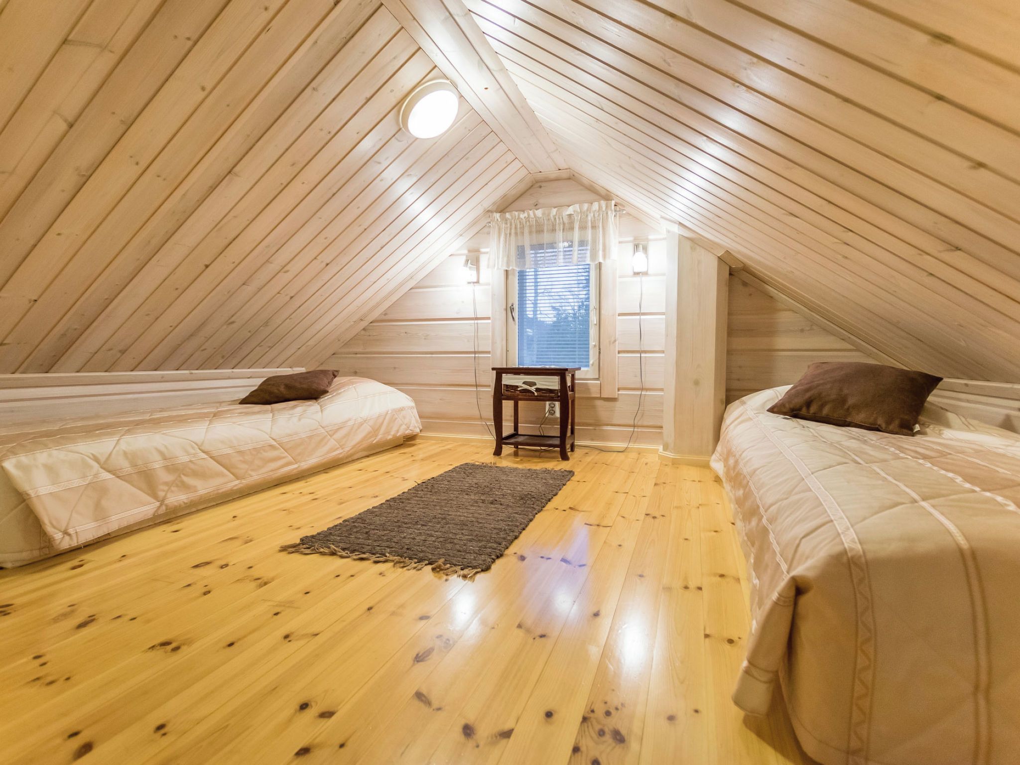 Um quarto de sótão pequeno e acolhedor, com paredes de madeira e teto inclinado. Há duas camas com almofadas e uma pequena mesa no centro.