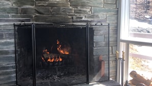 Une cheminée en pierre avec un feu de bois, une fenêtre donnant sur les arbres et un plancher en bois.
