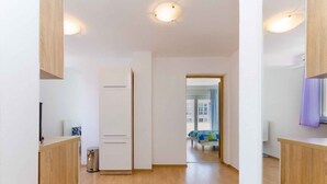 Un couloir moderne et épuré, avec un plancher en bois et des murs blancs. Une porte donne accès à une pièce où se trouve un lit et d’où l’on peut admirer la vue sur les bâtiments environnants.
