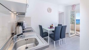 Une cuisine compacte équipée d’un évier, d’une cuisinière et d’un grille-pain. Une table à manger avec des chaises est placée près d’une porte-fenêtre coulissante qui donne sur un balcon.