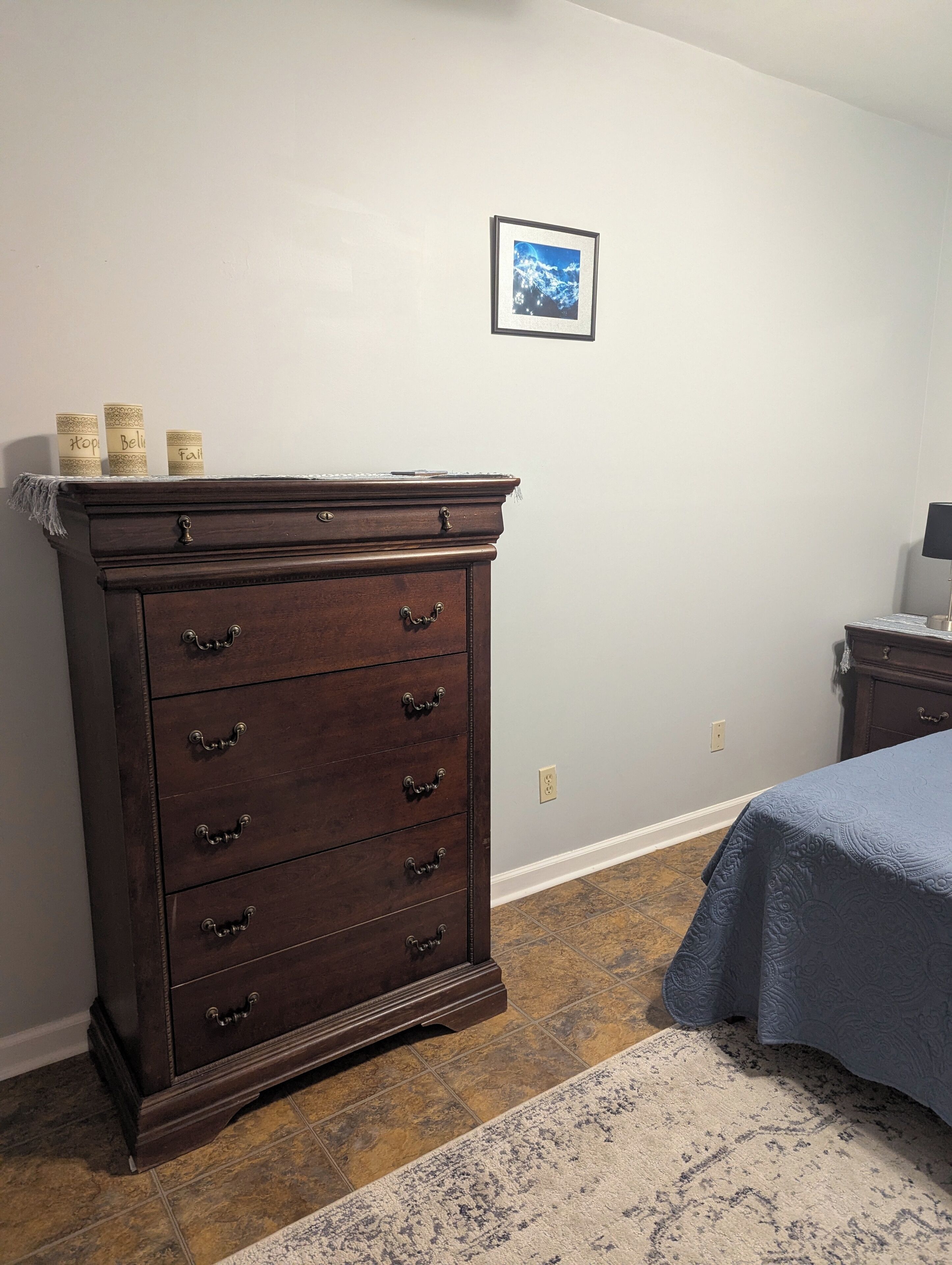 Une chambre avec une grande commode en bois, un lit recouvert d’une courtepointe bleue, une petite table avec une lampe et un tableau encadré accroché au mur.
