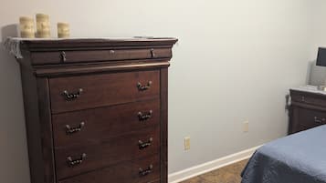 Une chambre avec une grande commode en bois, un lit recouvert d’une courtepointe bleue, une petite table avec une lampe et un tableau encadré accroché au mur.
