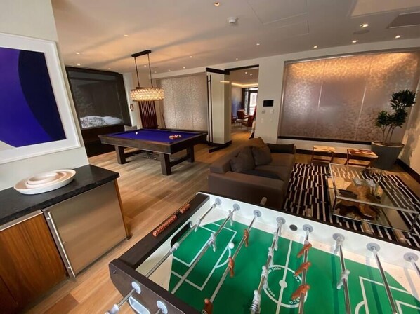 A modern living room with a pool table, a sofa, a foosball table, and a large window with patterned curtains.