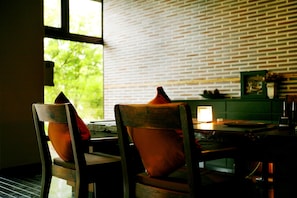 A dining area with brick walls, two wooden chairs with orange cushions, a table with a lamp, and a window with a view of greenery. - Tetsuka Ryokan (Satsuma)