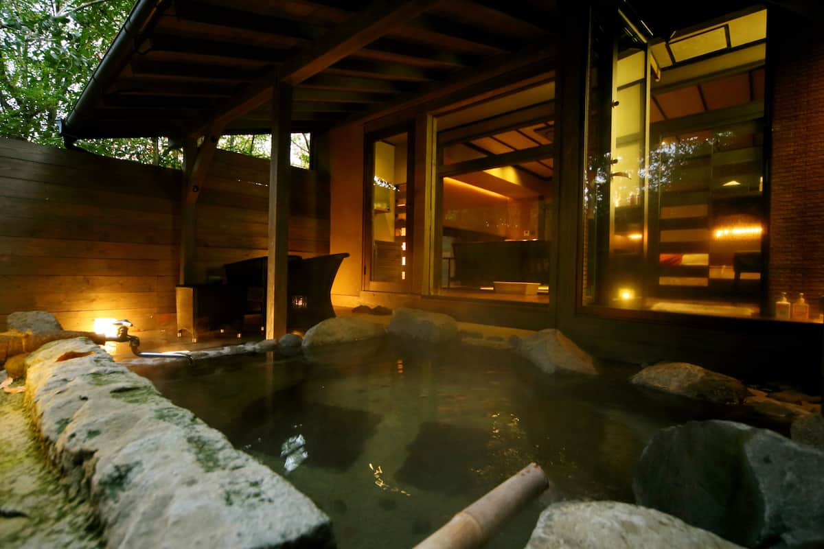Un espace de sources chaudes doté d’une terrasse en bois, d’un sentier en pierre et d’une porte en verre donnant accès à un espace intérieur.