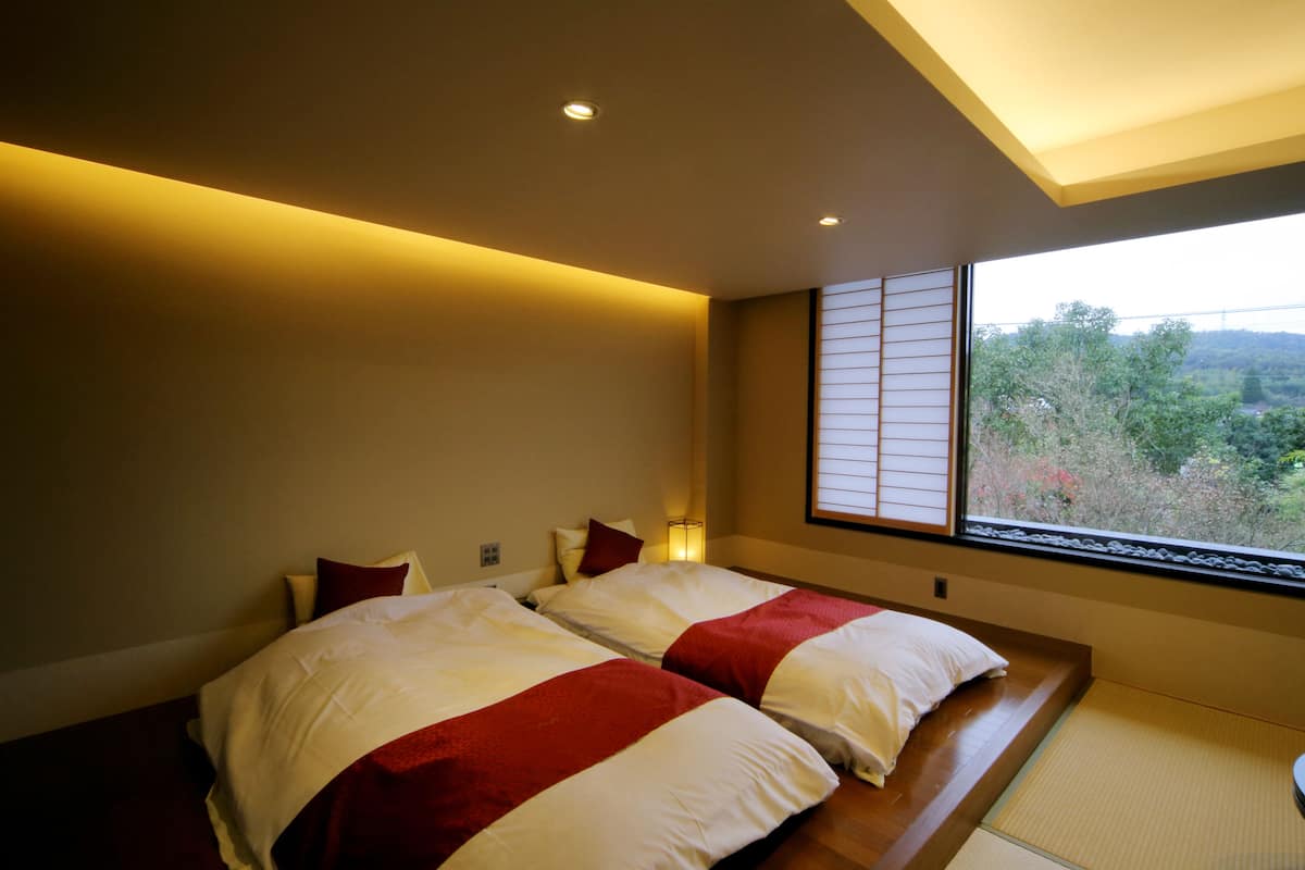 Une chambre d’hôtel moderne avec deux lits, une grande fenêtre donnant sur des arbres et des collines, et une porte coulissante de style japonais traditionnel.