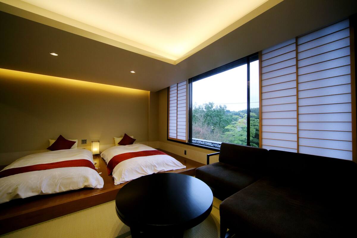 Une chambre d’hôtel avec deux lits, un canapé, une table ronde et une vue sur la verdure grâce à de grandes fenêtres.