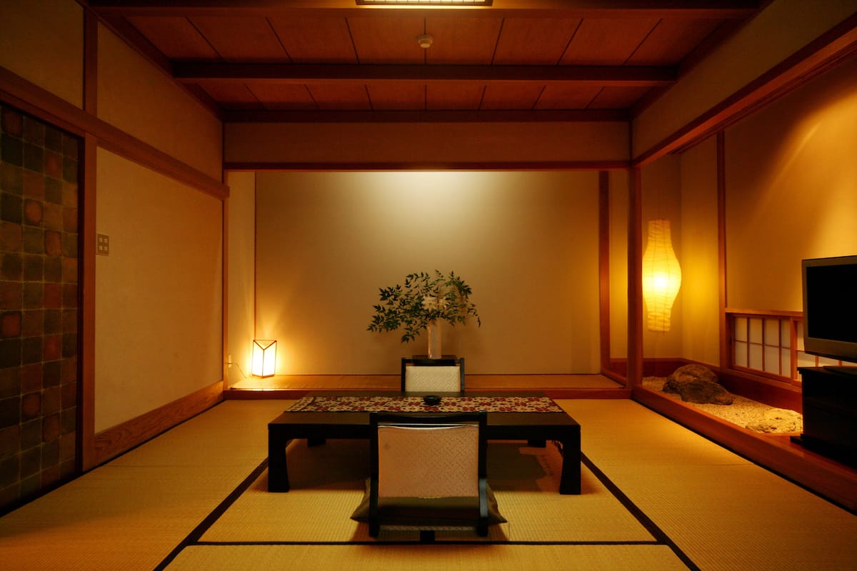 Une pièce de style japonais traditionnel, avec un sol recouvert de tatamis, une table basse, une chaise, une plante en pot et un téléviseur.