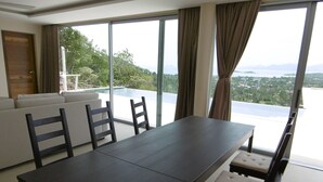 Un espace de repas moderne, avec une table et des chaises en bois foncé, de grandes fenêtres avec des rideaux et une vue sur un paysage luxuriant.