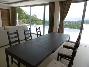 A modern dining area with a dark wooden table and chairs, large windows with curtains, and a view of a lush landscape.