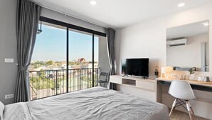 Habitación de hotel con una cama grande, un escritorio con televisor, una silla y un balcón con vistas a la ciudad.