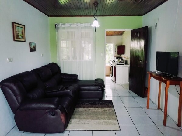 A living room with a sectional sofa, a television on a wooden stand, and a kitchen in the background.