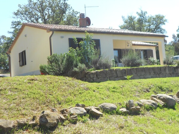 A single-story house with a tiled roof, a stone wall, and a grassy yard with scattered rocks.