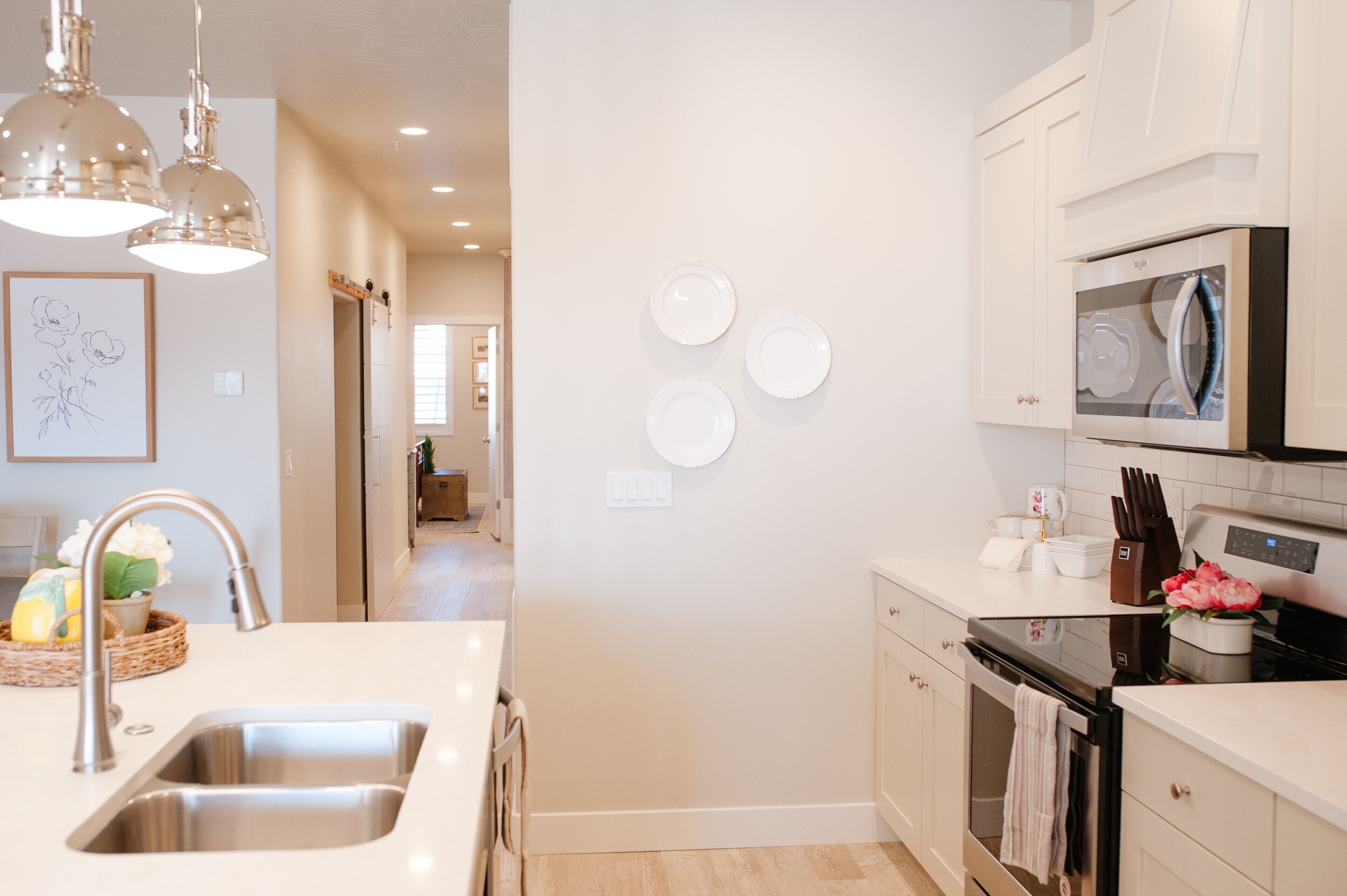 A modern kitchen with a stainless steel sink, white cabinets, a microwave, and a stove. There are three white plates on the wall and a framed picture above the sink.