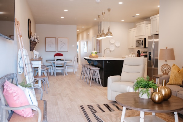Cocina moderna con armarios blancos, electrodomésticos de acero inoxidable y una isla central. Hay un comedor con mesa y sillas, y un área de descanso con sofá y mesa de centro.