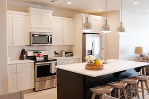 Una cocina moderna con gabinetes blancos, electrodomésticos de acero inoxidable, una isla central con taburetes e iluminación colgante.