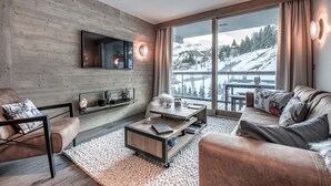 A modern living room with a wooden wall, a flat-screen TV, a sofa, a coffee table, and a view of a snowy landscape through large windows. - Winter Courchevel / Phoenix 502, proche commerce (Courchevel)