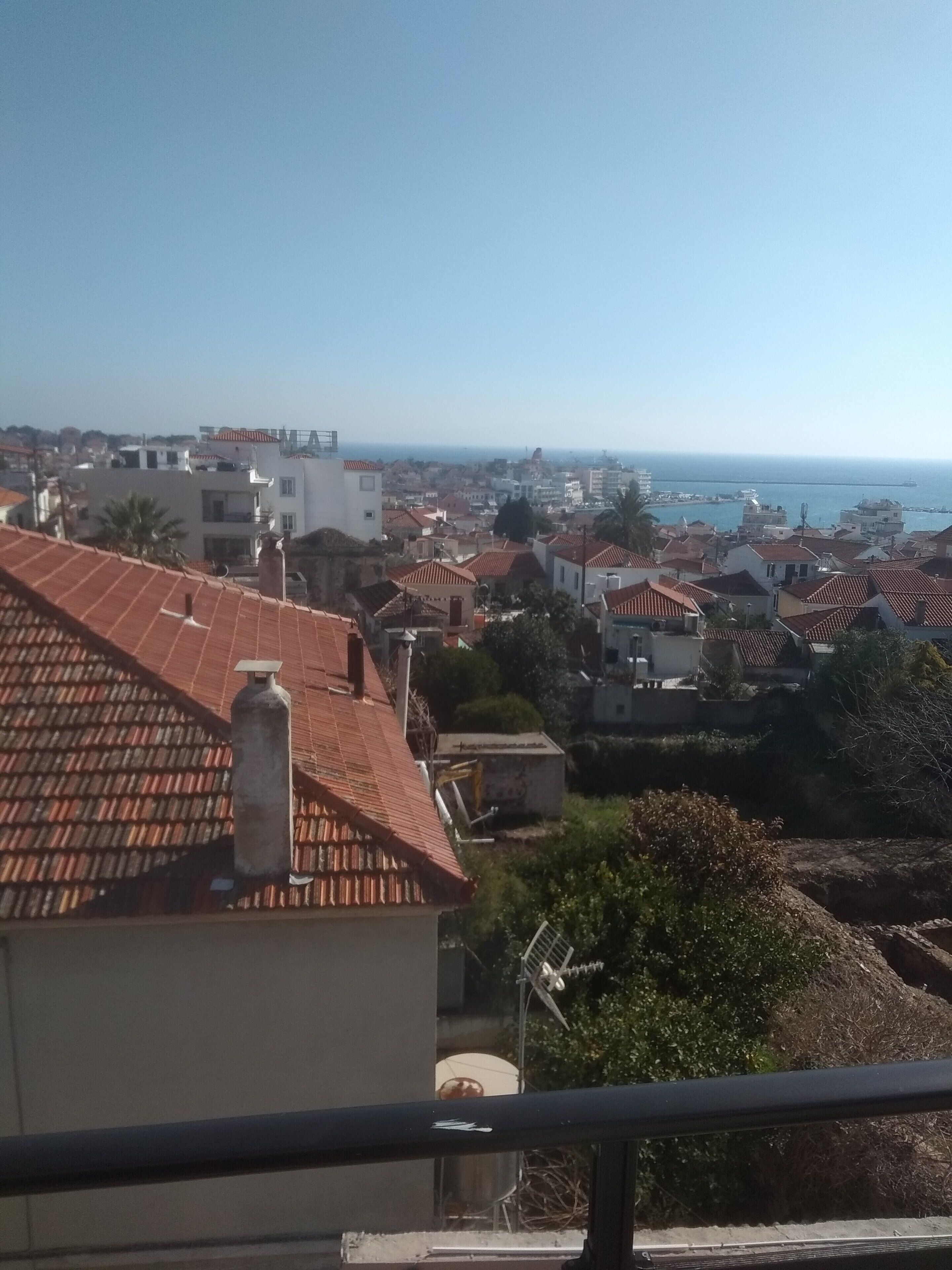 A coastal residential area with terracotta-tiled roofs, a clear sky, and a distant view of the sea.