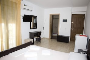 A hotel room with a bed, a desk, a chair, a small table, a TV, an air conditioner, a door, and a shower area. - Pyrgos Sofokleous (Lesvos)