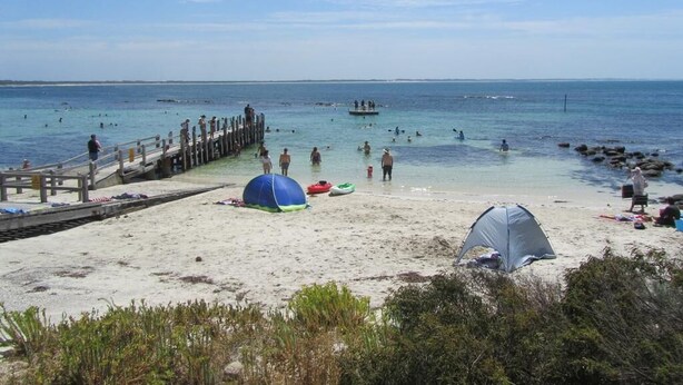 Una playa con un muelle de madera, una tienda de campaña y gente nadando en el agua.