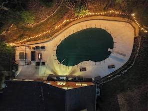 Vue aérienne d’un espace piscine avec une piscine à l’eau verte, une terrasse périphérique et des guirlandes lumineuses.
