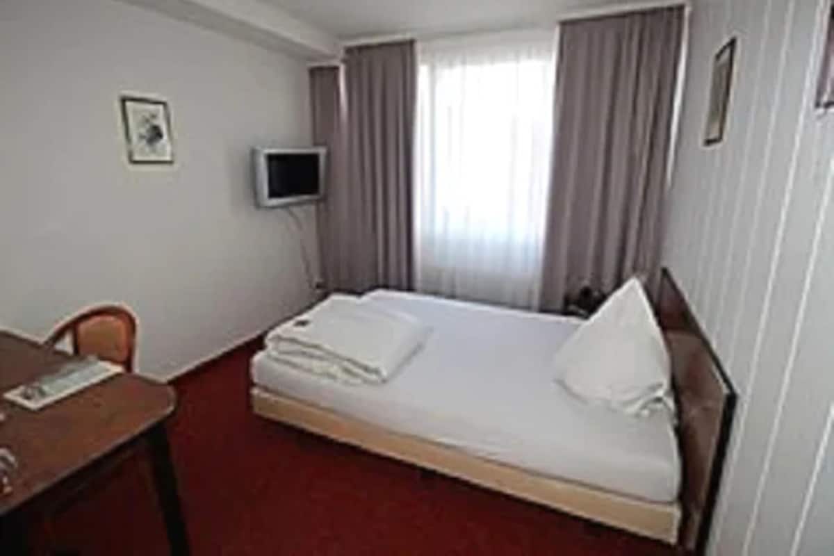 Habitación de hotel con una cama individual, un escritorio, una silla, un televisor y una ventana con cortinas.