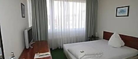 Una habitación de hotel con una cama, un escritorio con una computadora, una silla, una ventana con cortinas y un cuadro en la pared.