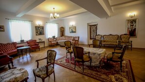 A spacious room with a high ceiling, wooden flooring, and a mix of seating including sofas and chairs. There are wall-mounted lamps and a chandelier. - Dwor Strzyzew (gmina Kampinos)