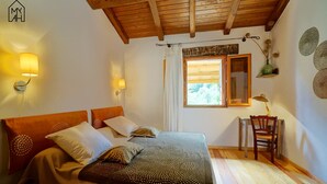A bedroom with a wooden ceiling, a bed with patterned bedding, a desk with a chair, and a window with curtains. - greenery in the chestnut grove. (Tasso)