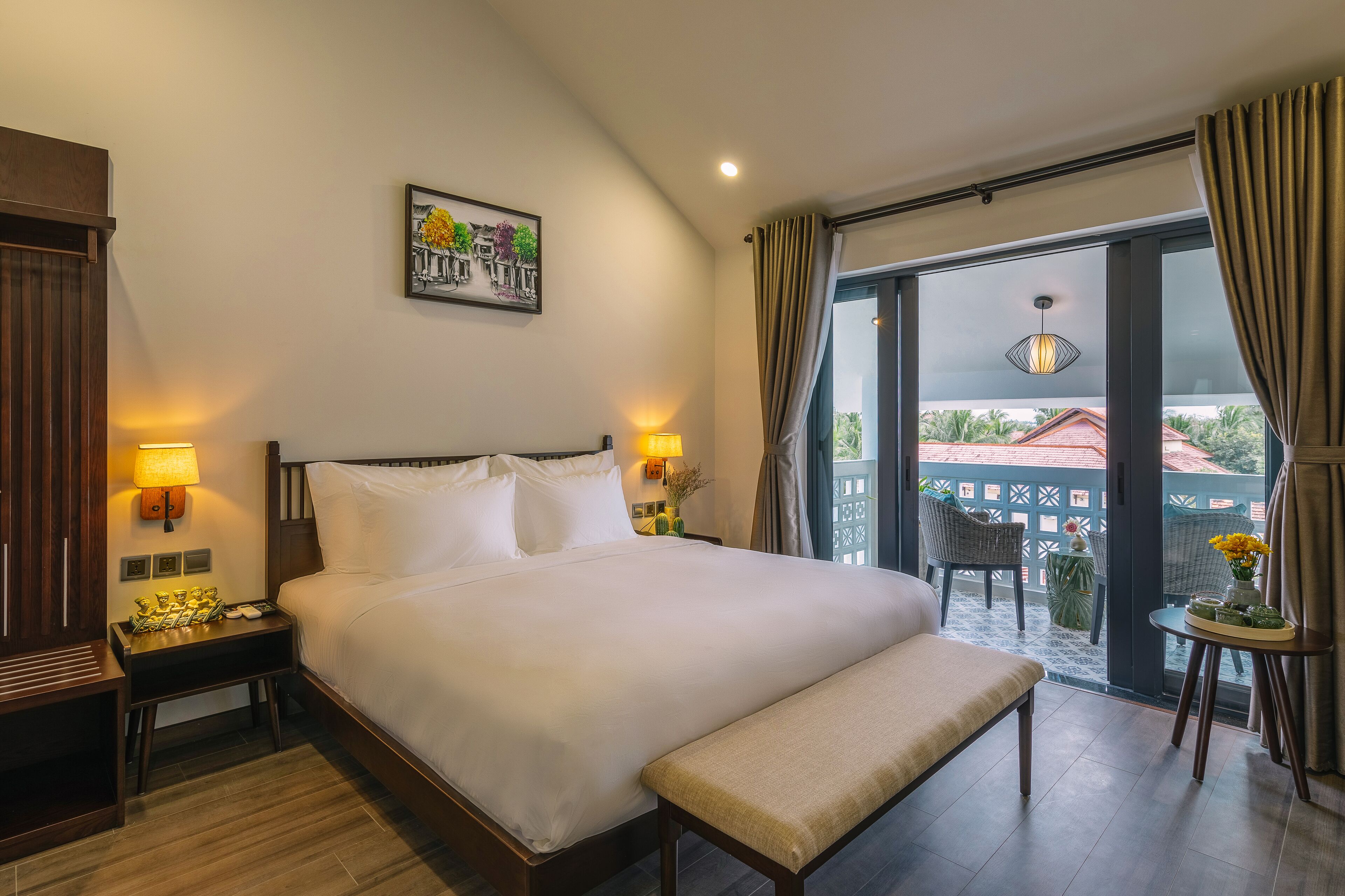 A hotel room with a large bed, bedside lamps, a bench, a small table with flowers, and a view of the outside through a glass door.