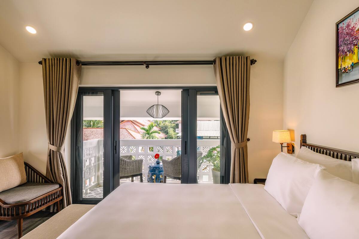 A hotel room with a large bed, two chairs, a nightstand with a lamp, and a view of a balcony with a railing and a potted plant.