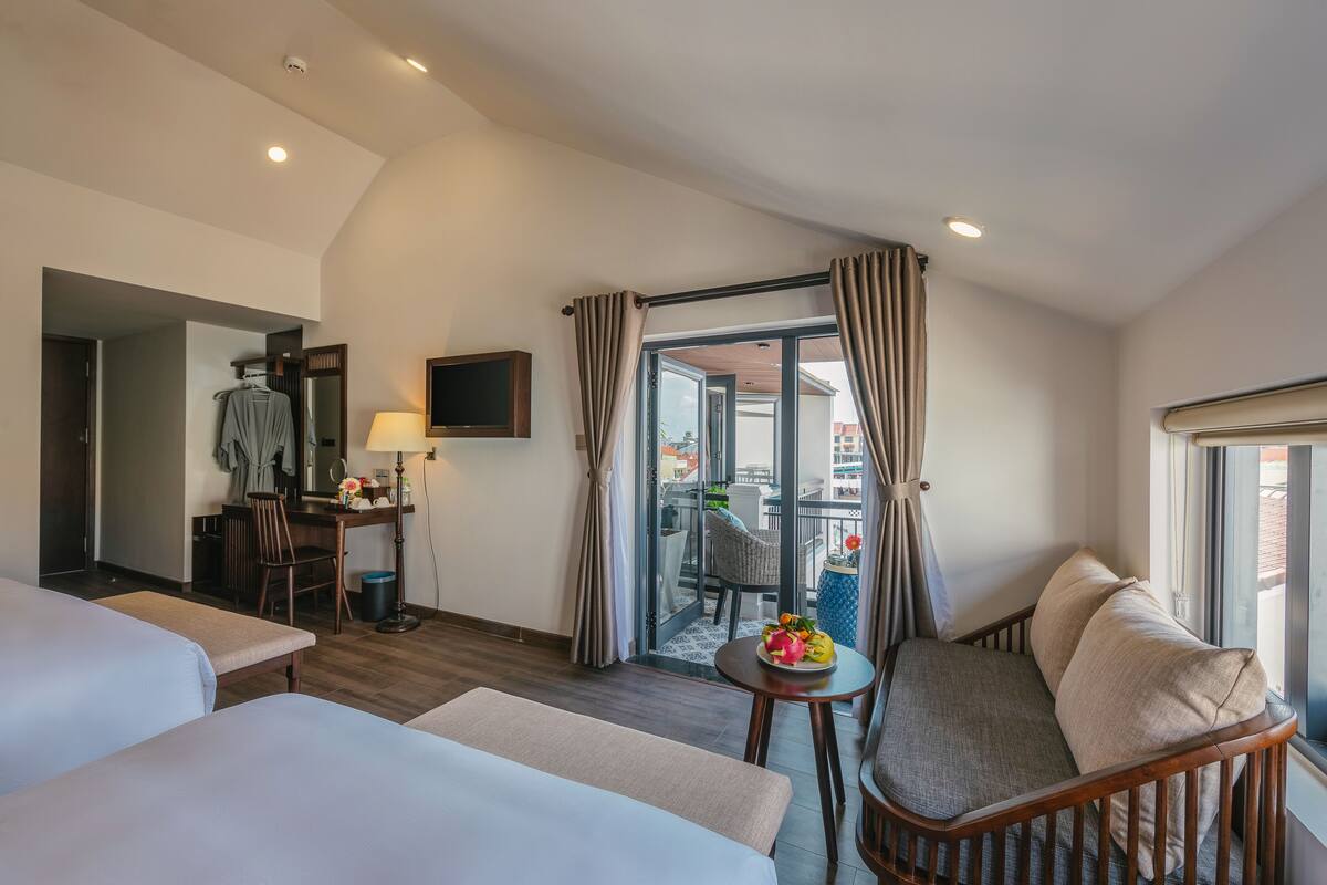 A hotel room with two beds, a desk with a chair, a small table with a fruit bowl, and a balcony with a view.