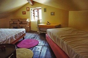 A small, cozy attic bedroom with two single beds, a wooden desk, a chair, and a window with a view of the outside.
