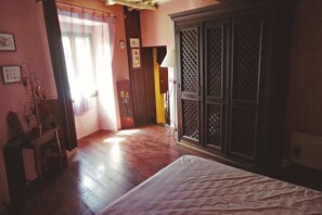 Una habitación con una cama, piso de madera, un armario, una mesa con lámpara y una ventana con cortinas.