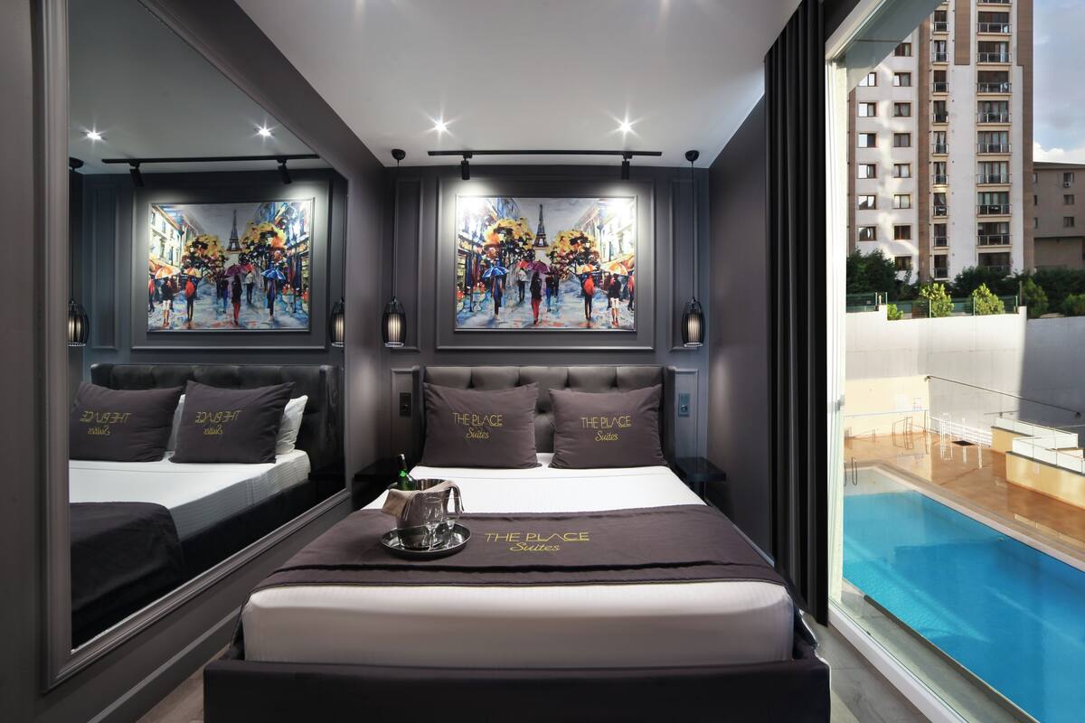 A hotel room with a large bed, a painting of a busy street scene, and a view of a swimming pool.