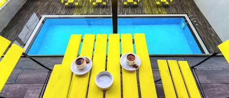 Zona de piscina con sillas reclinables amarillas y una mesa de vidrio, dos tazas de café y un cuenco blanco pequeño.