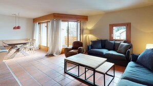 A living room with a sofa, a coffee table, a chair, and a dining table. There is a window with curtains and a lamp. - Very well located apartment house in the favorite and quiet Salet area. (St. Moritz)