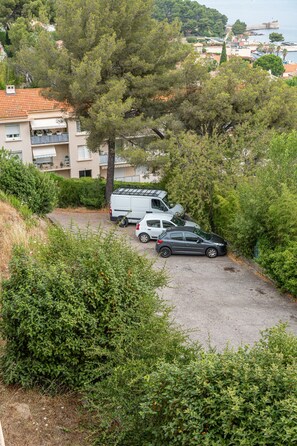 Ein Parkplatz mit einem weißen Lieferwagen und einem dunklen Auto, umgeben von dichter Vegetation und einem Gebäude mit Balkonen.