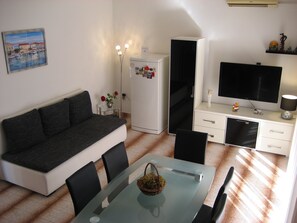 Ein kompakter Wohnraum mit einer Couch, Esstisch, Stühlen, einem Kühlschrank, einem Fernseher und einem kleinen Deko-Regal.