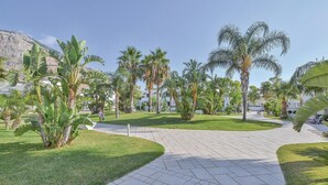 Ein gepflasterter Weg, umgeben von üppiger Vegetation und Palmen, führt zu einem Gebäude mit weißer Fassade.